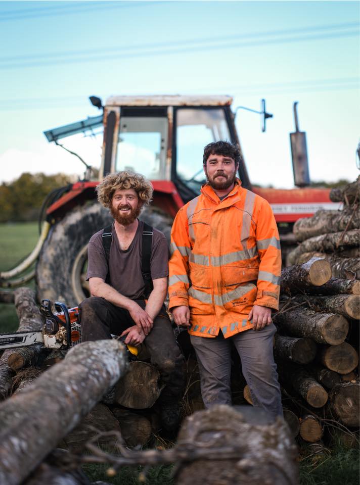 T & H Logs founders in front of their tractor and a stack of fresh-cut hardwood