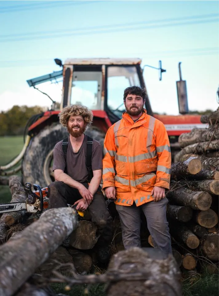 T & H Logs founders in front of their tractor and a stack of fresh-cut hardwood